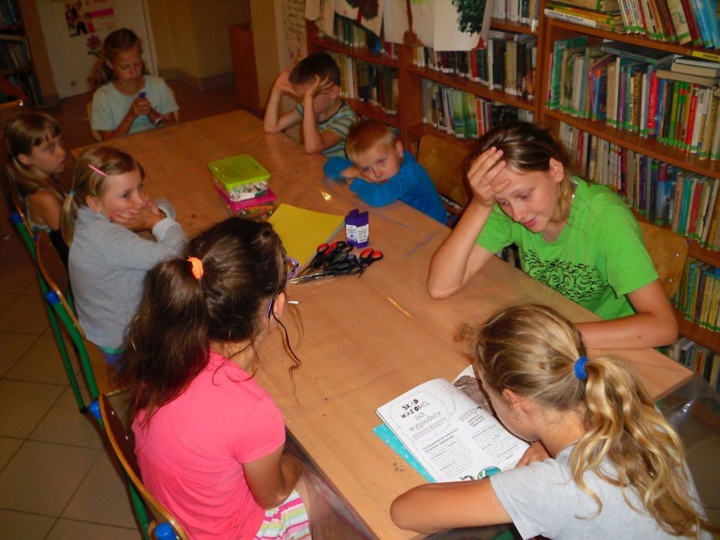 dzieci w czasie wakacyjnych zajęć w bibliotece słuchają opowiadania czytanego przez jedną z uczestniczek