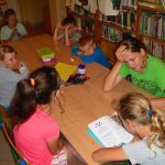 dzieci w czasie wakacyjnych zajęć w bibliotece słuchają opowiadania czytanego przez jedną z uczestniczek