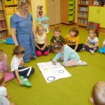 Pani bibliotekarka klęczy na podłodze, wokół niej dzieci siedzą. Jedna z dziewczynek maluje na kartce kółka.