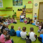 Dzieci z ochronki wraz z bibliotekarką siedzą na ziemi w kółku i patrzą co pokazuje pani bibliotekarka.