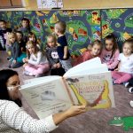 Grupa dzieci siedzi na podłodze. Pani bibliotekarka trzyma otworzoną książkę i pokazuje ilustracje.