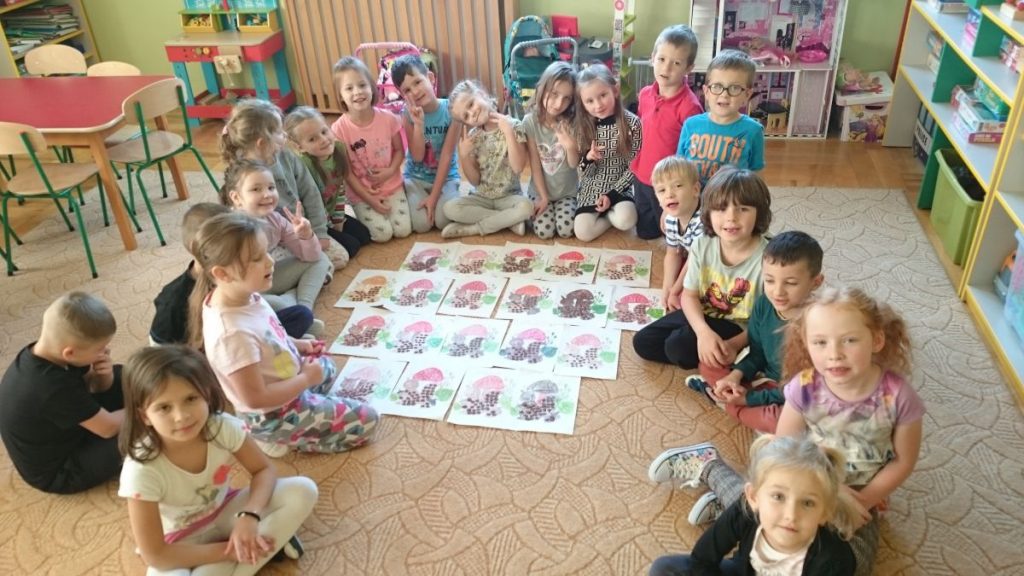 Przedszkolaki siedzące w kręgu prezentują wykonane przez siebie prace plastyczne z wizerunkami wiewiórek.