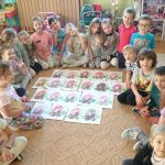 Przedszkolaki siedzące w kręgu prezentują wykonane przez siebie prace plastyczne z wizerunkami wiewiórek.