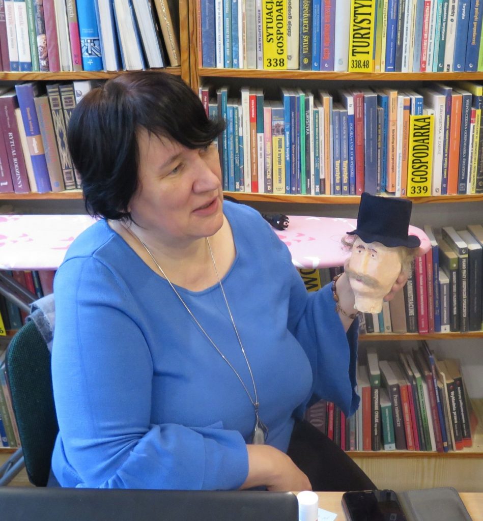 Na krześle siedzi pani dyrektor biblioteki Magdalena Krzeszowska trzyma w reku głowę manekina, który na głowie ma kapelusz.