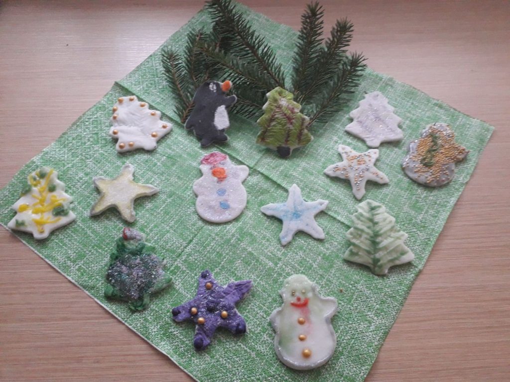 Na zdjęciu figurki wykonane z masy porcelanowej gwiazdki, mikołaj,choinka, aniołek. Połozone na zielonej serwetce wraz z zielonymi gałązkami .