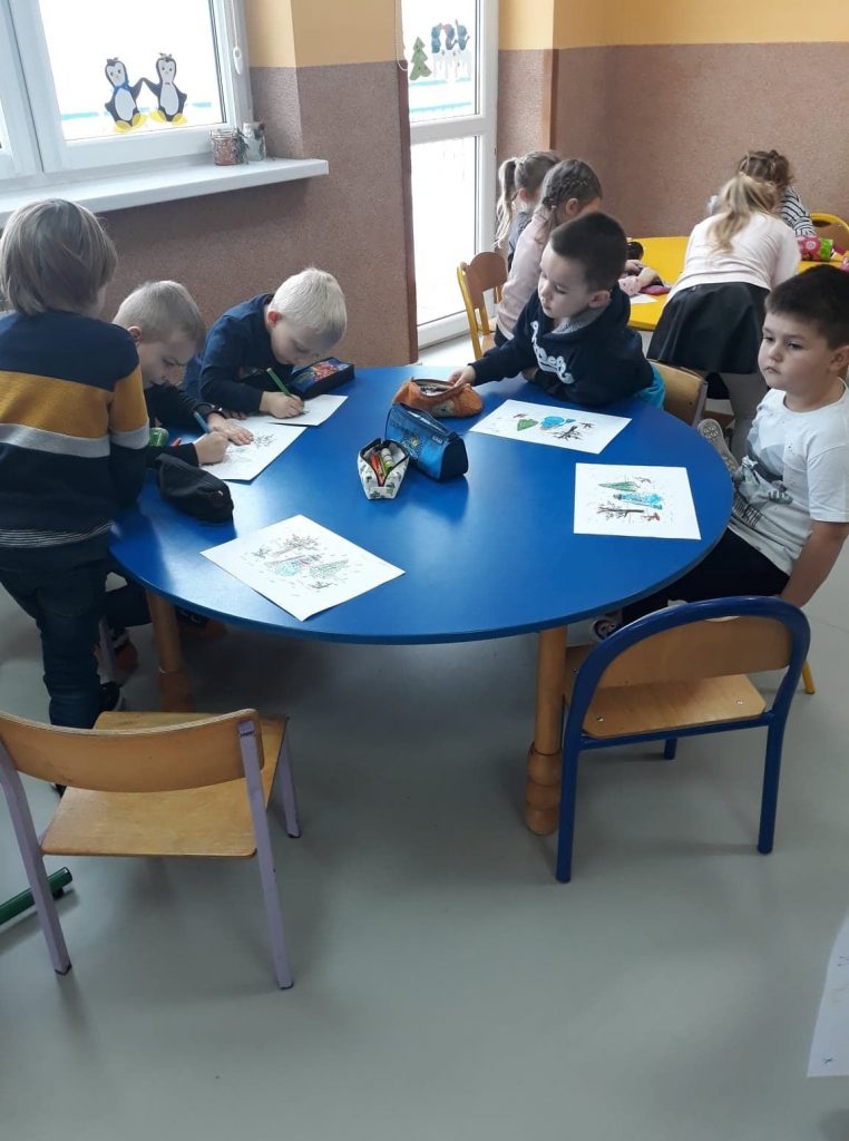 Grupa dzieci siedzi przy okrągłym stole i wykonuje prace plastyczne.
