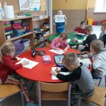 Grupa dzieci siedzi przy czerwonym i zielonym stole i wykonuje prace plastyczne.