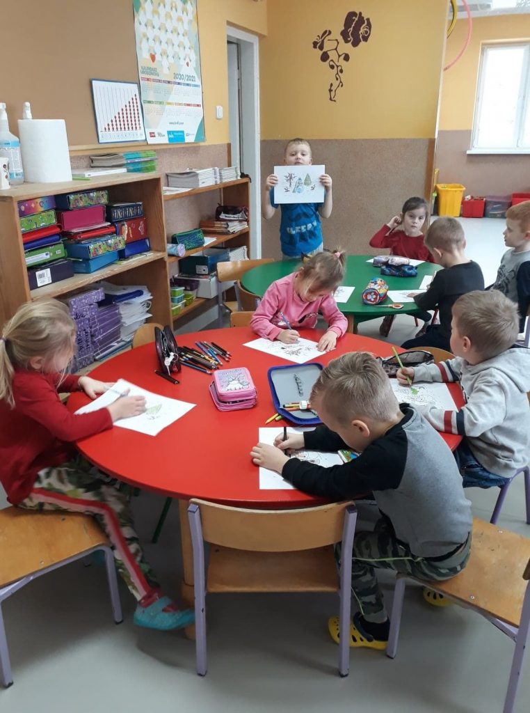 Grupa dzieci siedzi przy czerwonym i zielonym stole i wykonuje prace plastyczne.