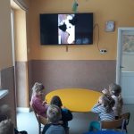 Grupa dzieci siedzi przy stole, i oglądaja bajkę w telewizorze powieszonym na ścianie, który powieszony jest na ścianie.