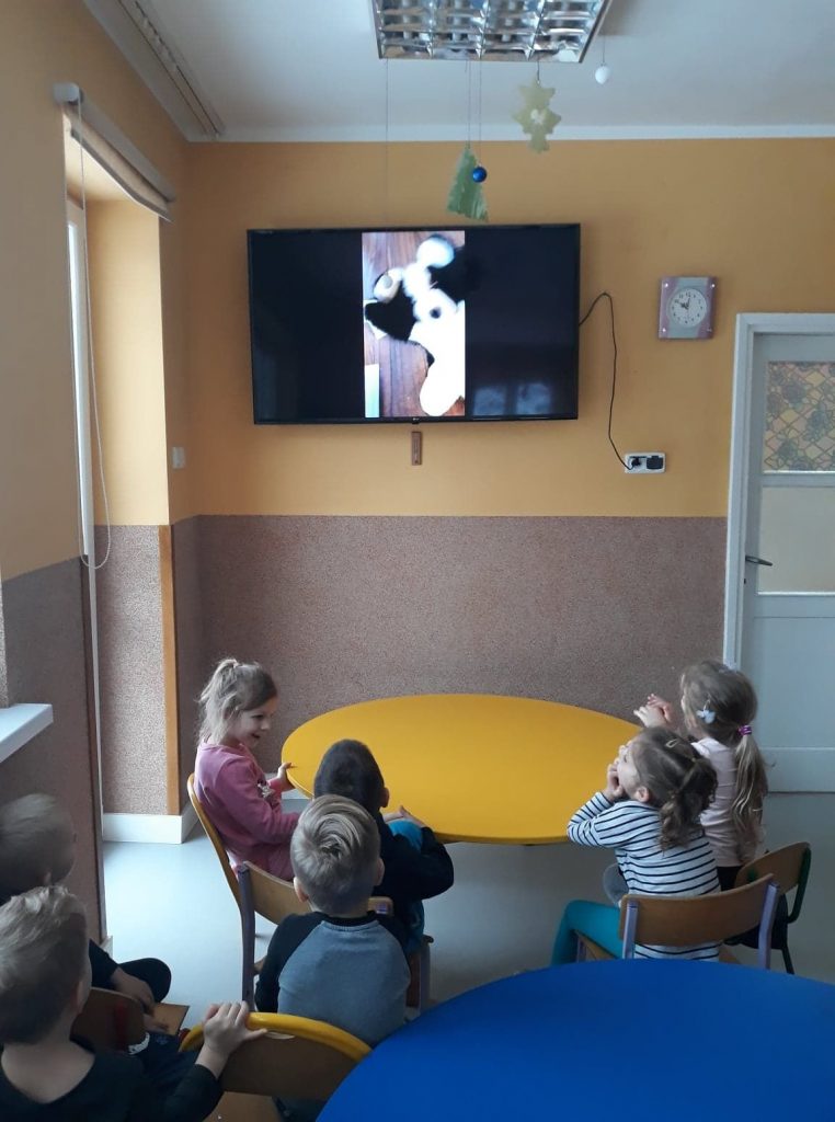 Grupa dzieci siedzi przy stole, i oglądaja bajkę w telewizorze powieszonym na ścianie, który powieszony jest na ścianie.