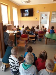 Spora grupa przedszkolaków siedzi na krzesełkach oraz na kocykach i ogląda z zaciekawieniem na zawieszonym na ścianie wielkim telewizorze film, na którym Pan Borsuk czyta im bajkę pt. „Być jak Tygrys”. 