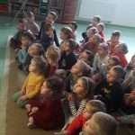 Na zdjęciu widać grupę przedszkolaków siedzących na jasnym dywanie, słuchających i oglądających na ekranie telewizora czytaną przez bibliotekarza bajkę pt. „Franklin przygotowuje święta”. W tle widoczne są regały z książkami oraz pięć obrazków w zielonych ramkach przedstawiających 4 krasnoludki i królewnę Śnieżkę.