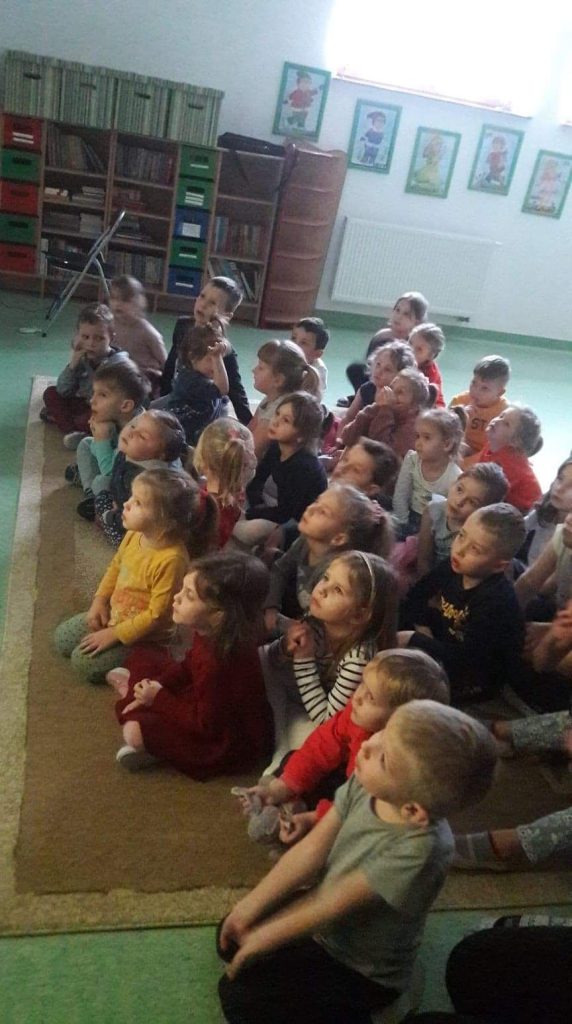 Na zdjęciu widać grupę przedszkolaków siedzących na jasnym dywanie, słuchających i oglądających na ekranie telewizora czytaną przez bibliotekarza bajkę pt. „Franklin przygotowuje święta”. W tle widoczne są regały z książkami oraz pięć obrazków w zielonych ramkach przedstawiających 4 krasnoludki i królewnę Śnieżkę.