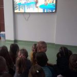 Na zdjęciu widać grupkę przedszkolaków słuchających i oglądających na ekranie telewizora czytaną przez bibliotekarza bajkę pt. „Franklin przygotowuje święta”.