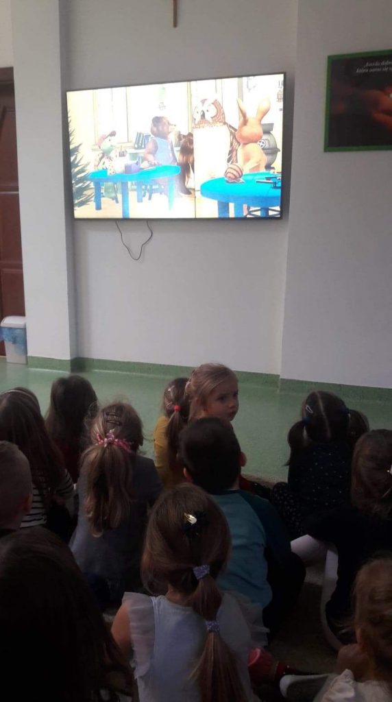 Na zdjęciu widać grupkę przedszkolaków słuchających i oglądających na ekranie telewizora czytaną przez bibliotekarza bajkę pt. „Franklin przygotowuje święta”.