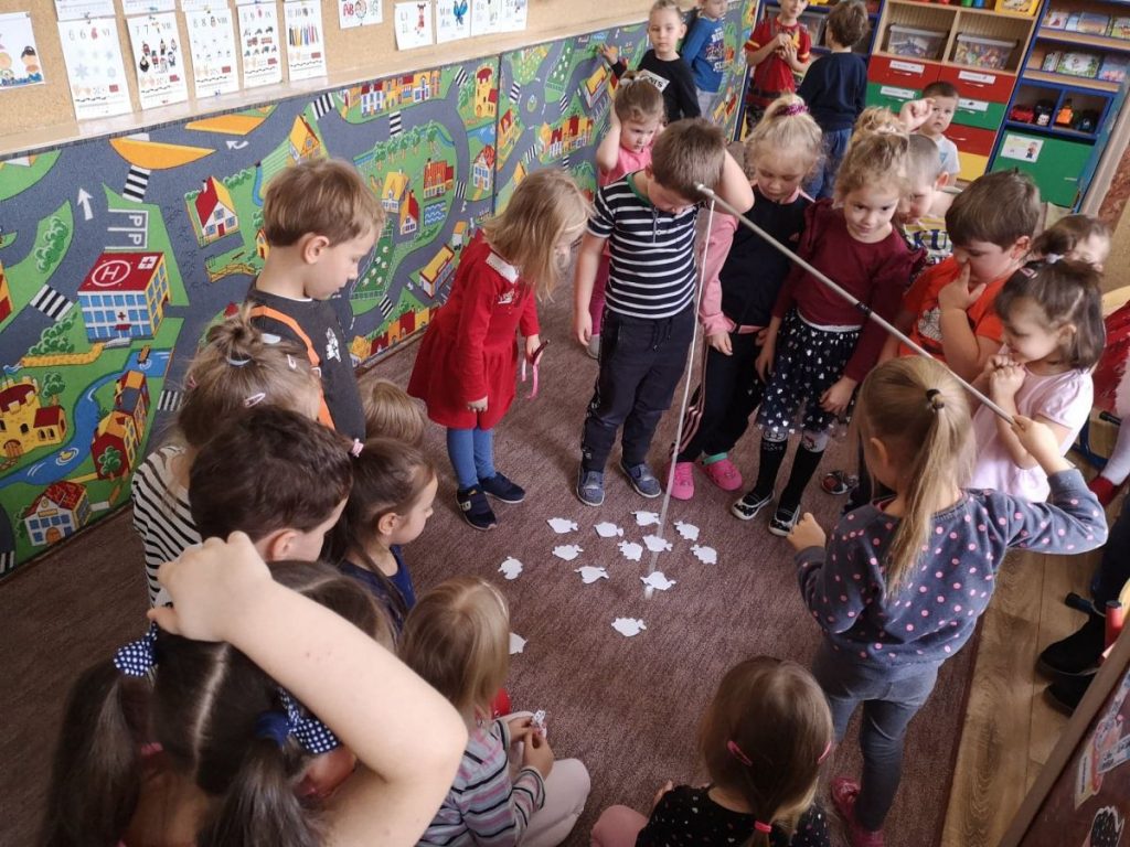 Dzieci stoją w sali przedszkolu w kółku. Jedna dziewczynka trzyma w tęku wędkę i łowi na papierowe rybki, ktore rosłożone sa na ziemi. Reszta dzieci obserwuje.