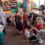 Pani bibliotekarka siedzi na małym krzesełku i czyta bajkę. Wokół niej dzieci siedzą na ziemi i słuchaja bajki.