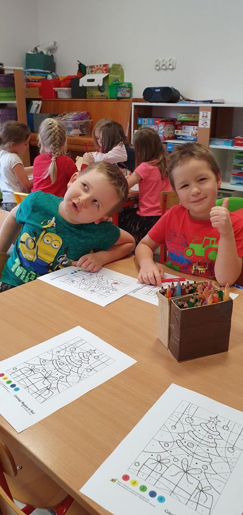 W sali przedszkola, przy stolikach siedzi dwóch chłopców z jednej grup przedszkolnych . Na stole położone kredki oraz kartki, które kolorują.