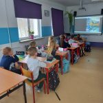 Grupa dzieci siedzi w ławkach i ogląda bajkę.