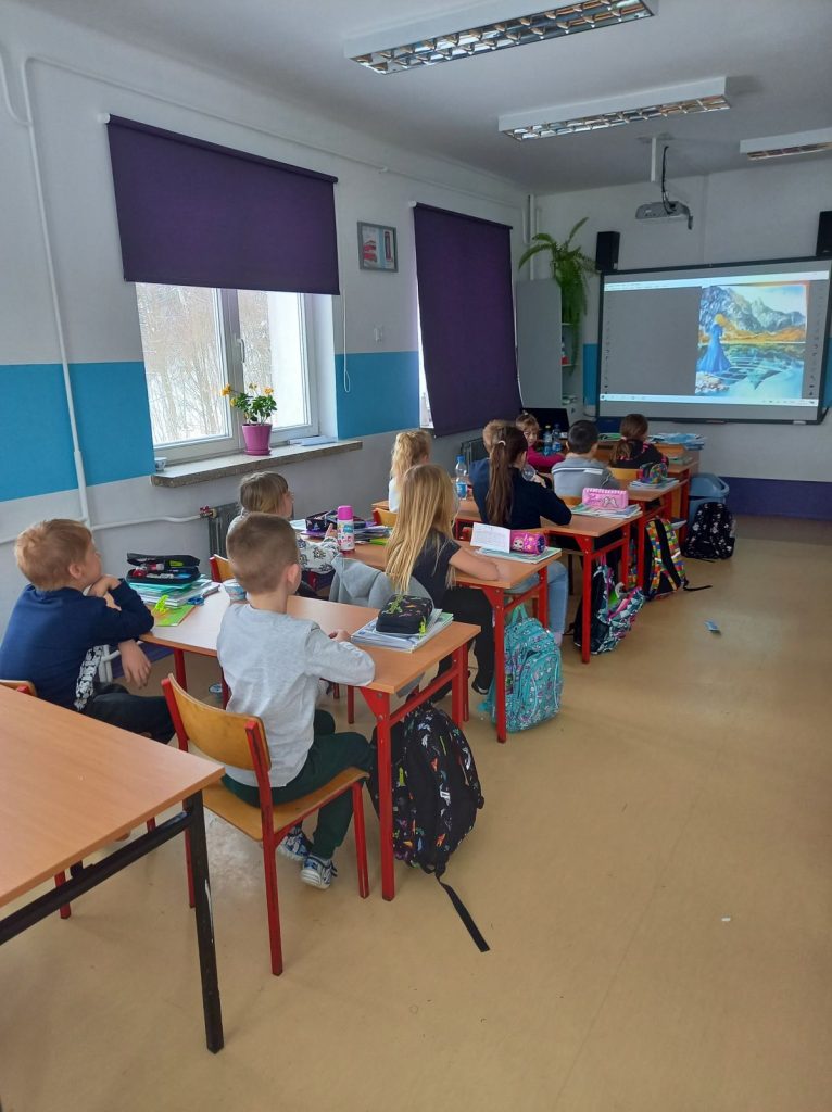 Grupa dzieci siedzi w ławkach i ogląda bajkę.