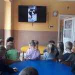 W przedszkolnej sali przedszkolaki siedząc na krzesełkach oglądają na ekranie telewizora czytającego im książkę Pana Borsuka