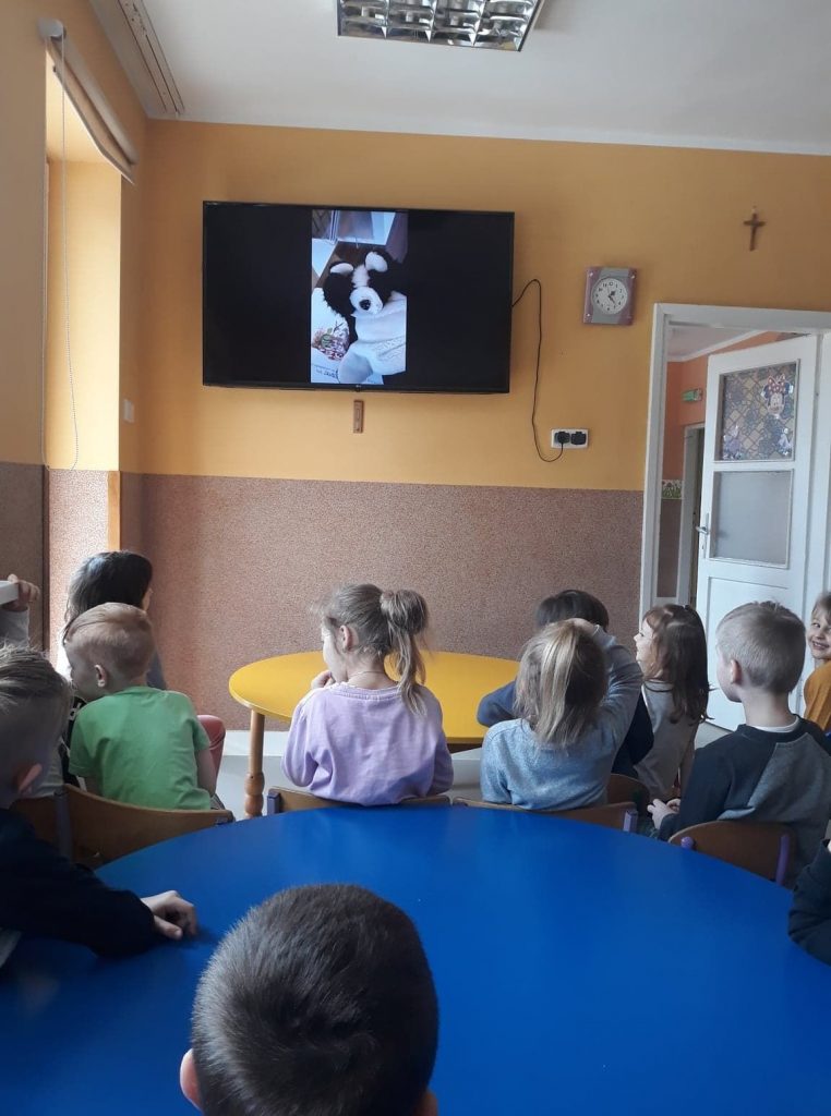 W przedszkolnej sali przedszkolaki siedząc na krzesełkach oglądają na ekranie telewizora czytającego im książkę Pana Borsuka