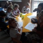 Przy małym żółtym stoliku siedzi grupa uśmiechniętych dzieci, każde koloruje kredkami sprytną leśną wiewiórkę