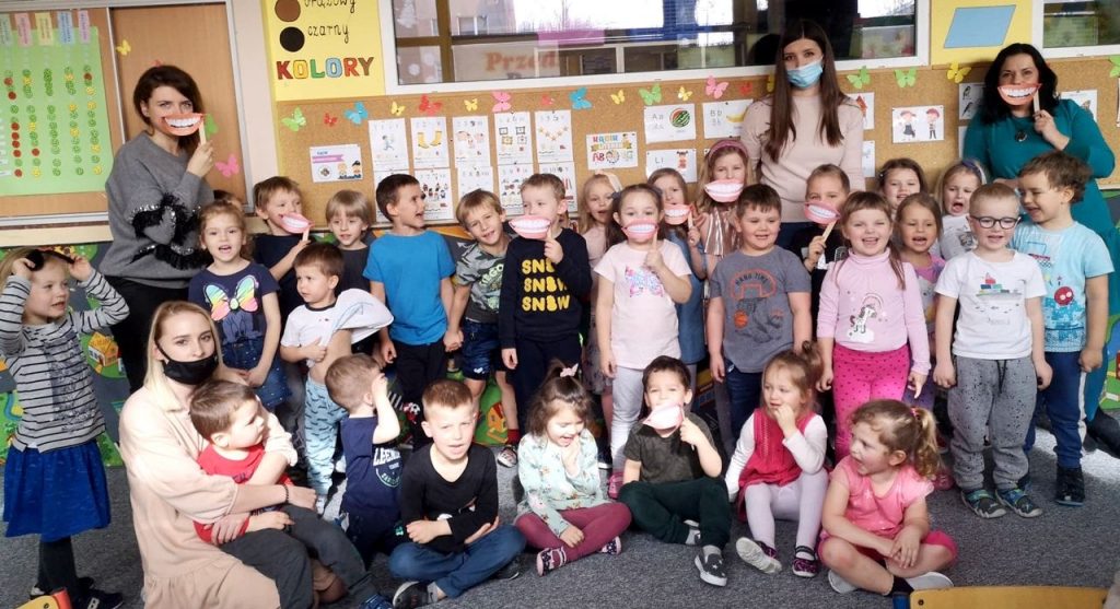 4. Zdjęcie grupowe na którym widać 4 panie, dwie z nich przyłożone mają do ust papierowe uśmiechy. Trzy panie stoją a jedna siedzi na podłodze trzymając na kolanach dziecko. Na fotografii widać około 30 przedszkolaków, są to dziewczynki i chłopcy. Część z nich stoi a część siedzi po turecku. Większość z nich jest uśmiechniętych, niektóre maja również papierowe uśmiechy. Widać również kolorowe dekoracje, obrazki, plakaty i tablice umieszczone na ścianach.