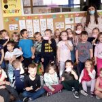4. Zdjęcie grupowe na którym widać 4 panie, dwie z nich przyłożone mają do ust papierowe uśmiechy. Trzy panie stoją a jedna siedzi na podłodze trzymając na kolanach dziecko. Na fotografii widać około 30 przedszkolaków, są to dziewczynki i chłopcy. Część z nich stoi a część siedzi po turecku. Większość z nich jest uśmiechniętych, niektóre maja również papierowe uśmiechy. Widać również kolorowe dekoracje, obrazki, plakaty i tablice umieszczone na ścianach.