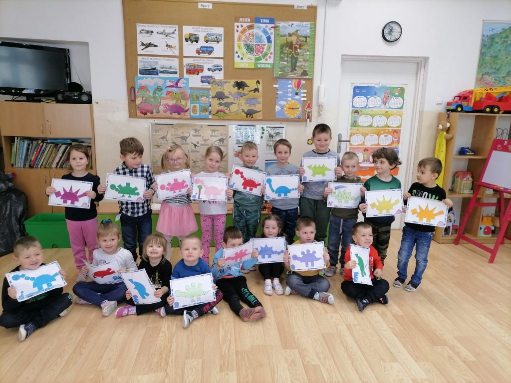 Na zdjęciu widoczna jest grupa przedszkolaków. Ośmioro przedszkolaków siedzi na podłodze, a dziesięcioro stoi w rzędzie za nimi. Dzieci pozują do zdjęcia z wykonanymi przez siebie pracami plastycznymi z wizerunkami dinozaurów powstałych z figur geometrycznych (kół, trójkątów, kwadratów, prostokątów). Powstałe prace plastyczne są w różnych kolorach: różowym, fioletowym, niebieskim, czerwonym, zielonym, pomarańczowym, żółtym. W tle za przedszkolakami widoczny jest telewizor stojący na regale z książkami, tablica z planszami o dinozaurach, pojazdach i porach roku oraz drzwi do Sali i regał z zabawkami.