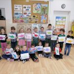 Na zdjęciu widoczna jest grupa przedszkolaków. Ośmioro przedszkolaków siedzi na podłodze, a dziesięcioro stoi w rzędzie za nimi. Dzieci pozują do zdjęcia z wykonanymi przez siebie pracami plastycznymi z wizerunkami dinozaurów powstałych z figur geometrycznych (kół, trójkątów, kwadratów, prostokątów). Powstałe prace plastyczne są w różnych kolorach: różowym, fioletowym, niebieskim, czerwonym, zielonym, pomarańczowym, żółtym. W tle za przedszkolakami widoczny jest telewizor stojący na regale z książkami, tablica z planszami o dinozaurach, pojazdach i porach roku oraz drzwi do Sali i regał z zabawkami.