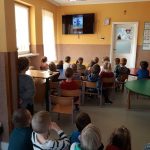 W przedszkolnej sali wszystkie zgromadzone dzieci z zainteresowaniem oglądają na wiszącym na ścianie wielkim telewizorze bajkę pt. „Papierowe Samoloty”