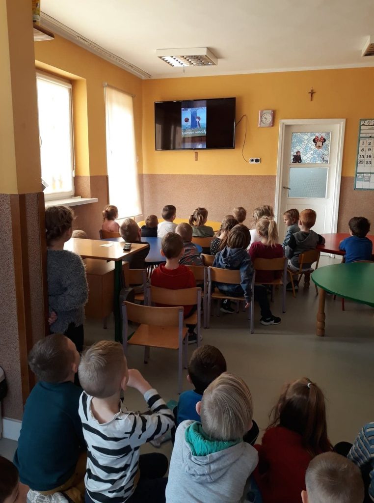 W przedszkolnej sali wszystkie zgromadzone dzieci z zainteresowaniem oglądają na wiszącym na ścianie wielkim telewizorze bajkę pt. „Papierowe Samoloty”