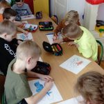 Przy długim stole siedzą dziewczynki i chłopcy i z zapałem rysują kredkami na kartkach papieru kolorowe samoloty