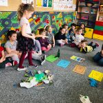 Na zdjęciu widać dzieci w wieku przedszkolnym siedzące na popielatej wykładzinie. Jedna dziewczynka stoi i trzyma w ręku tekturowe pudełko. Na podłodze rozrzucane są różne śmieci i przedmioty, które przedszkolaki mają za zadanie posegregować do koszy w odpowiednim kolorze wydrukowanych na kartkach. Dzieci znajdują się w sali przedszkolnej, w tle widać korowe szafki na których znajdują się zabawki. Na ścianach przybita jest kolorowa wykładzina a powyżej znajdują się różnego rodzaju tablice z literkami, zwierzętami itp.tematyczne.