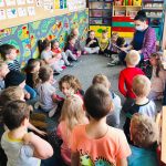 Zdjęcie przedstawia dzieci siedzące na ziemi w kole. Są to dziewczynki i chłopcy w wieku przedszkolnym. W kole na małym krzesełku siedzi bibliotekarka czytająca im książkę. Bibliotekarka posiada na buzi maseczkę. Wszyscy znajdują się w sali przedszkolnej. W tle widać korowe szafki na których znajdują się zabawki.