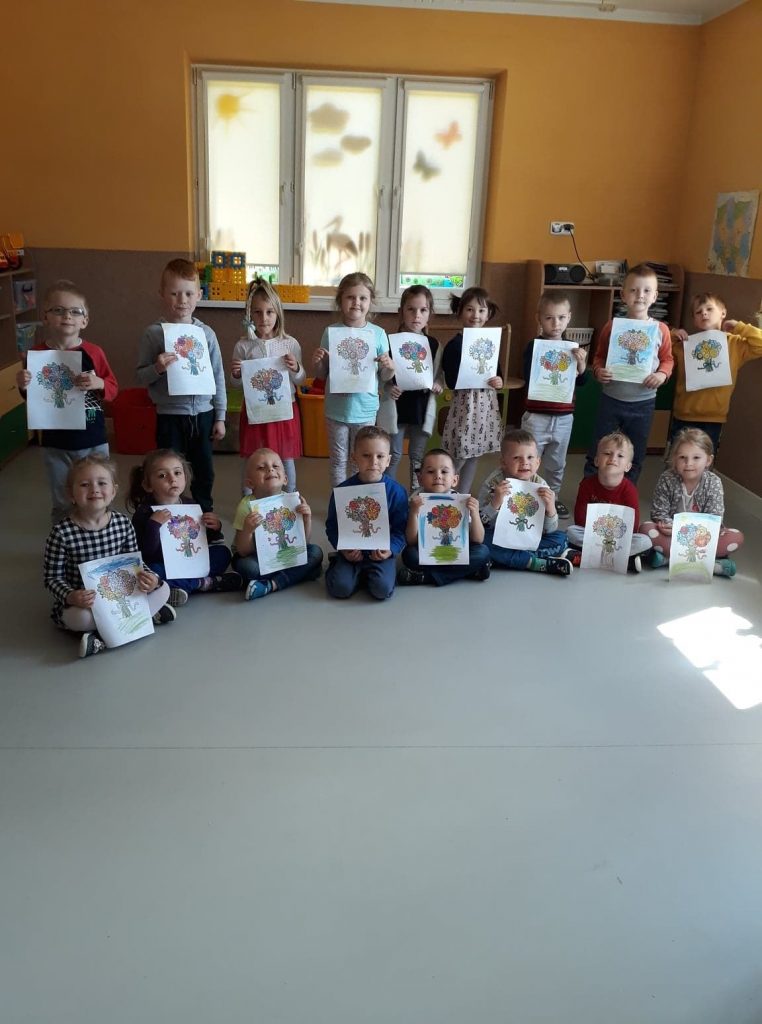 Grupa przedszkolaków ustawiła się do grupowego zdjęcia, każde z dzieci trzyma w rączkach namalowany przed momentem piękny rysunek.