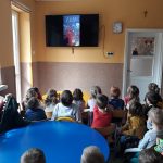 Przedszkolaki siedzą na krzesełkach i z zadartymi głowami oglądają wyświetlaną im na wiszącym na ścianie monitorze bajkę pt. „Drzewo”