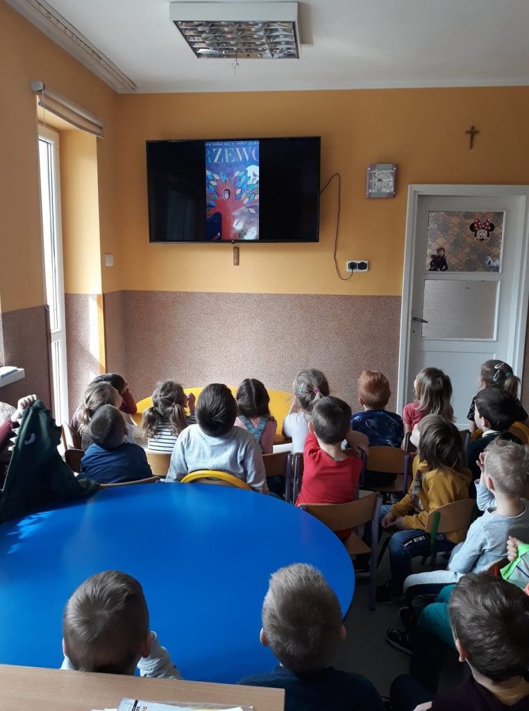 Przedszkolaki siedzą na krzesełkach i z zadartymi głowami oglądają wyświetlaną im na wiszącym na ścianie monitorze bajkę pt. „Drzewo”