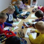 Przy stoliku siedzą obok siebie dzieci, które w skupieniu rysują kredkami na kartkach kolorowe drzewa