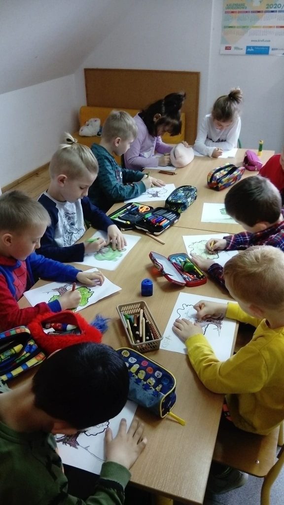 Przy stoliku siedzą obok siebie dzieci, które w skupieniu rysują kredkami na kartkach kolorowe drzewa