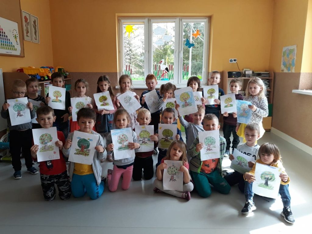 Jedna z grup przedszkolaków ustawiła się pod oknem do pamiątkowego zdjęcia, uśmiechnięte dzieci trzymają w rączkach swoje rysunki