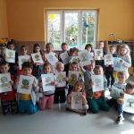 Jedna z grup przedszkolaków ustawiła się pod oknem do pamiątkowego zdjęcia, uśmiechnięte dzieci trzymają w rączkach swoje rysunki