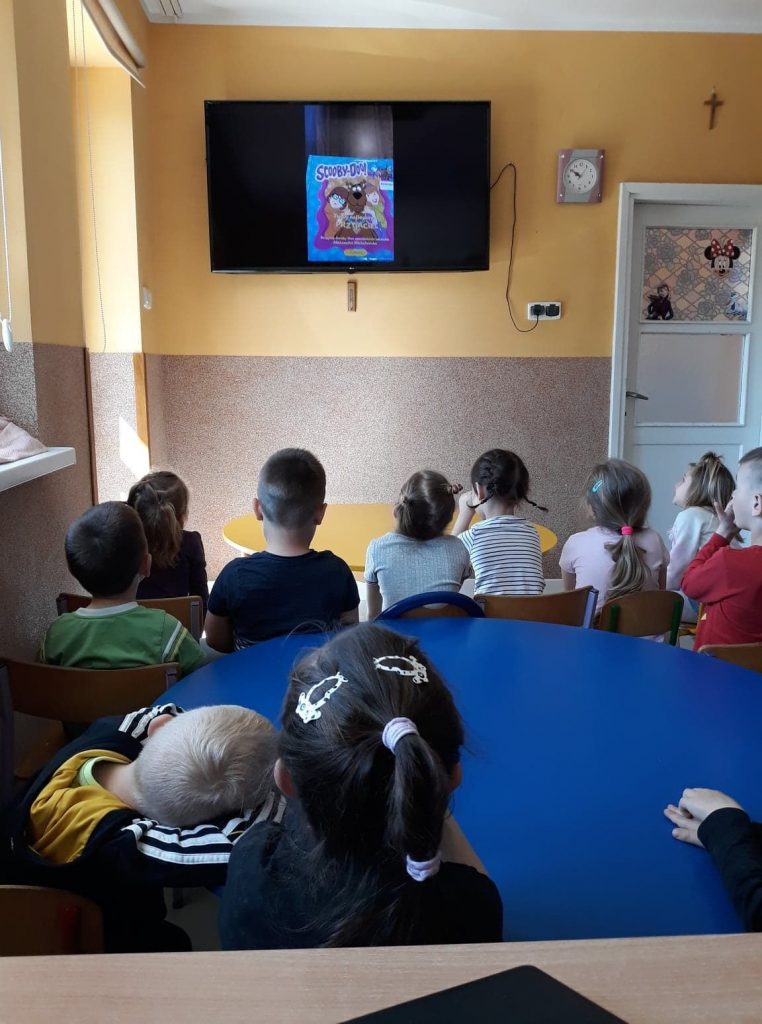 W sali przedszkolnej grupa dzieci słucha czytanej im przez Pana Borsuka bajki o śmiesznym Scooby-Doo