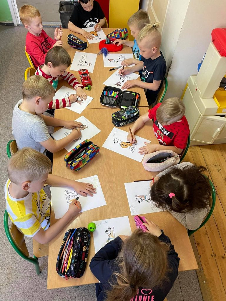 Przedszkolaki siedzą przy długim, zapełnionym pomocami plastycznymi stoliku i w skupieniu rysują kredkami na kartkach portrety bajkowych psiaków