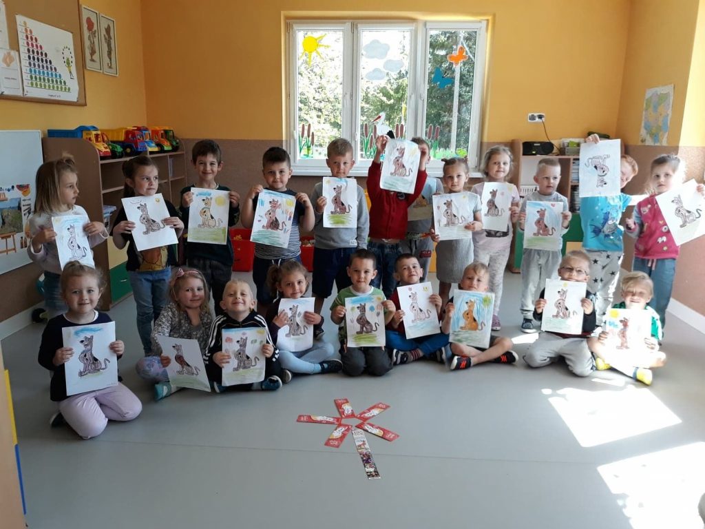 Grupa przedszkolaków ustawiła się pod oknem do wspólnej fotografii, dzieci trzymają w rękach swoje kolorowe obrazki, a na podłodze leży ułożony z zakładek kwiatek