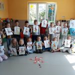 Grupa przedszkolaków ustawiła się pod oknem do wspólnej fotografii, dzieci trzymają w rękach swoje kolorowe obrazki, a na podłodze leży ułożony z zakładek kwiatek