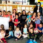 Zdjęcie przedstawia grypę dzieci w w wieku przedszkolany. Są to zarówno dziewczynki i chłopcy . Część dzieci siedzi, część stoi, z tyłu w drugim rzędzie widać również trzy Panie. Dwoje dzieci trzyma książki a kilka duży rysunek przedstawiający rodzinę.