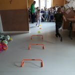 W dużej przedszkolnej sali rozłożono tor przeszkód składający się z niskich płotków i kolorowych stożków, zadaniem dzieci jest pokonać go jak najsprawniej.