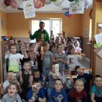 Liczna grupa uśmiechniętych przedszkolaków pozuje razem z bibliotekarzem trzymając w rękach pamiątkowe kolorowe zakładki.